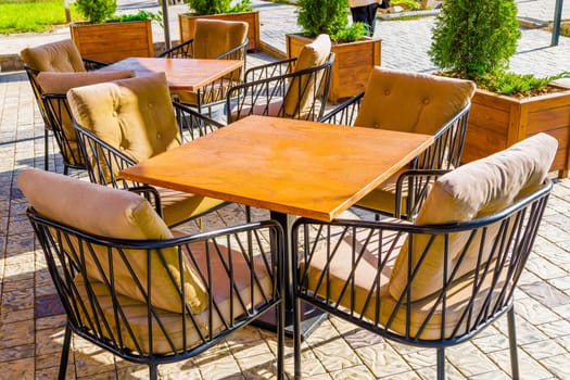 The charming outdoor seating area features plush cushions on steel chairs and lovely wooden tables in a bright, sunlit ambiance, no people
