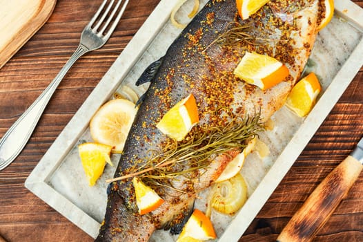 Delicious roasted trout seasoned with spices and orange on a stone serving board on wooden background. Close up.