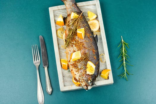Delicious roasted trout seasoned with spices and orange on a stone serving board on wooden background. Close up.