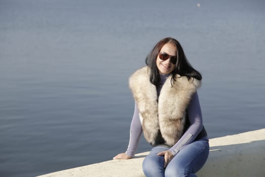 A stylish woman is smiling by the serene water, wearing trendy sunglasses and a cozy fur vest to enhance her look