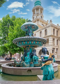 Ganga, Azerbaijan - May 13 2024: Fountain with sculptures, located outside the Ganja State Philharmonic Hall. The large, ornate building has a light-colored facade, with green trees along the sidewalk