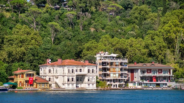 In Istanbul, Turkey, on a summer day, one can admire the scenic view of the lush green mountains and traditional houses nestled among dense trees on the Asian side of the Bosphorus strait