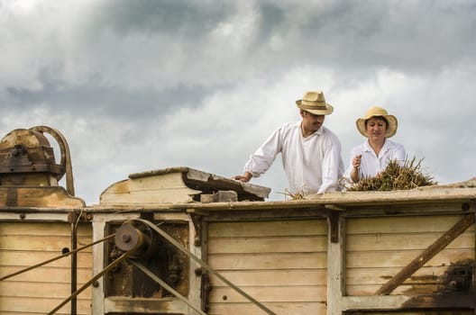 Pleudihen-sur-Rance, France - August 12, 2007: Two French farmers using ancient machine, threshing grain by removing seeds from the stalks and husks of hay on ancient combine harvester