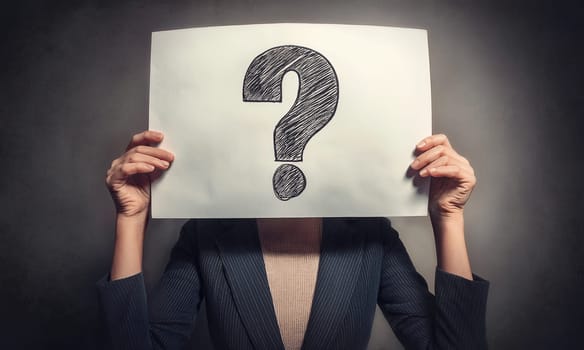 Businesswoman covers face using a sheet with drawn question mark isolated over dark concrete wall background. Woman business person with a paper page hiding face identity like a mask