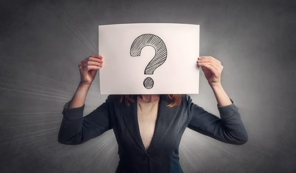 Businesswoman covers head using a paper sheet with drawn question mark isolated on dark wall background. Woman hiding face identity like a mask, feel stressed, emotional discomfort and anxiety