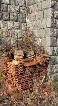 A pile of weathered terracotta roof tiles sits at the base of a stone wall, with dried grass and debris suggesting abandonment or disuse.
