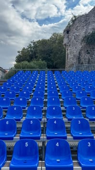 Rows of blue plastic numbered seats in an outdoor seating area, backed by trees and rock formations