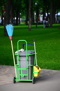 Cleaning service cart full of supplies and equipment along with grey trash bin for professional cleaner against green grass in public park