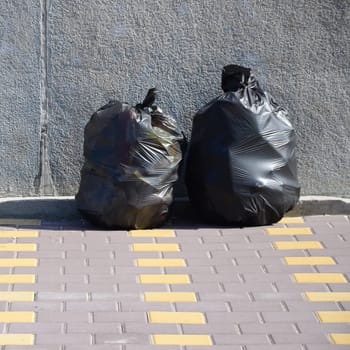 Two black garbage bags on tiled street floor at concrete fence in city. Pollution trash