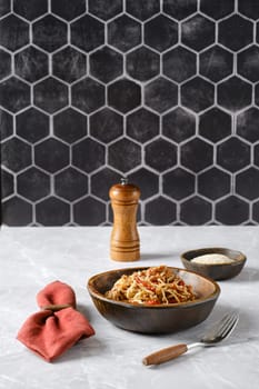 A bowl of spaghetti with pulled beef topped with fresh herbs and spices on a marble countertop, accompanied by a pepper grinder and a small dish of grated cheese