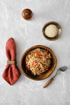 A bowl of stir-fried noodles and pulled beef with red peppers and garnished with sesame seeds rests on a marble countertop. A wooden bowl of sesame seeds and a red napkin accompany it.