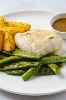 A succulent piece of white fish is plated alongside vibrant green beans and crispy roasted potatoes, accompanied by a small bowl of savory sauce for dipping.