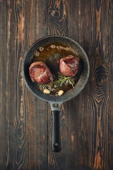 Two beef fillets are sizzling in a skillet with garlic and rosemary, showcasing the cooking process on a rustic wooden countertop. The aroma of the meat fills the kitchen with warmth and flavor.