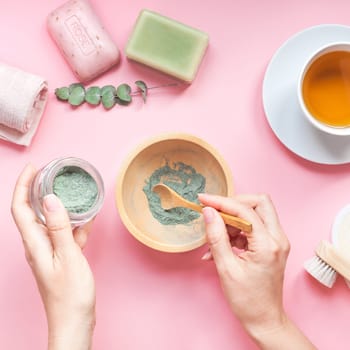 Spa, beauty cosmetics and body care treatment concept copy space. Creative top view flat lay of woman hands with bath accessories, green spirulina cleansing mask in bowl, cup of tea pink background