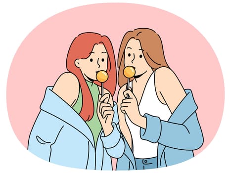 Two women with chupa chups in hands dressed in fashionable seductive clothes eat lollipops. Lovely girls in stylish jackets slung off shoulders to advertise new candies for teenagers