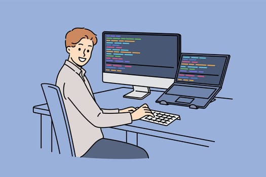 Man programmer types code and looks at camera, sitting at table with computer and laptop. Successful guy programmer develops software or application, working in team with UI or UX designers