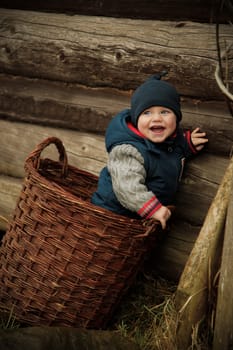 A cheerful child beams widely with a bright smile while seated in a wicker basket by a rustic wooden wall