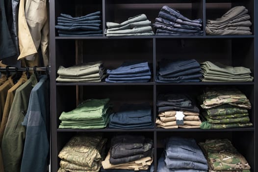 Stacked tactical pants and military uniforms, including camouflage and outdoor gear, neatly arranged on dark wooden shelves in a store.