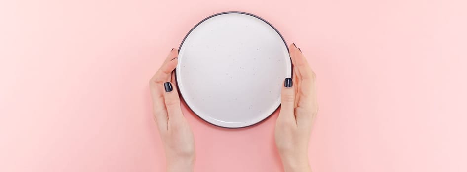 Creative flat lay top view of woman hands black manicure with empty plate mock up on pastel millennial pink paper background copy space. Minimal dish service concept template for your text menu design