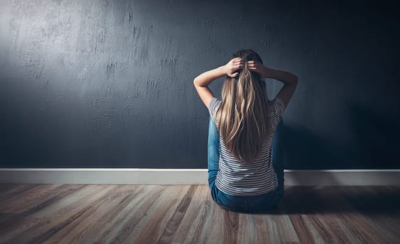 Rear view stressed teenage girl seated on the floor in a dark room, looking hopeless in front of the wall, keeps hands to head. Sad woman feels emotional discomfort, anxiety and mental health problems
