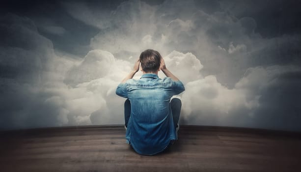 Rear view stressed male sitting on the floor hands to head, looking hopeless in front of the wall with clouds texture. Pessimistic guy with head in the clouds feels anxiety and mental health problems