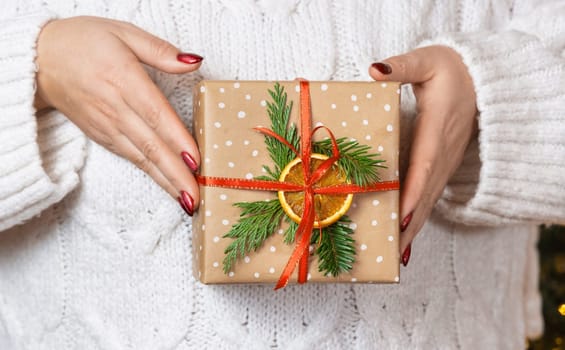 Woman in white knitted sweater with Christmas gift in hands, front view. Girl with red nails with wrapped and decorated present. Cozy holidays atmosphere