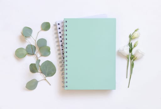 Light green spiral notebook on a white table near green eucalyptus twigs and flowers top view, Blank textbook mockup. Minimal school flat lay.