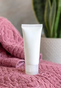White cream squeeze tube on pink folded towels near green plant in bathroom, close up, cosmetic packaging mockup. Natural mens face cream for personal care