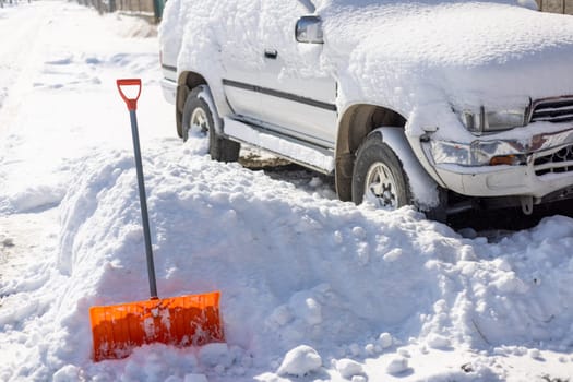 An orange snow shovel sticks out of a snowdrift in front of a snow-covered white SUV car on a sunny winter day