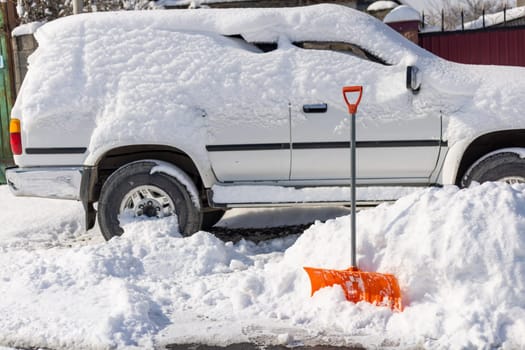 An orange snow shovel sticks out of a snowdrift in front of a snow-covered white SUV car on a sunny winter day