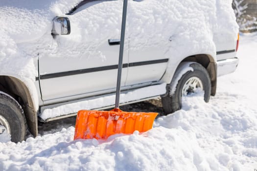 An orange snow shovel sticks out of a snowdrift in front of a snow-covered white SUV car on a sunny winter day