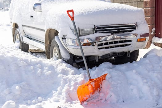 An orange snow shovel sticks out of a snowdrift in front of a snow-covered white SUV car on a sunny winter day