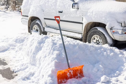 An orange snow shovel sticks out of a snowdrift in front of a snow-covered white SUV car on a sunny winter day