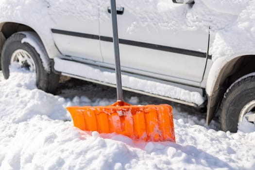 An orange snow shovel sticks out of a snowdrift in front of a snow-covered white SUV car on a sunny winter day