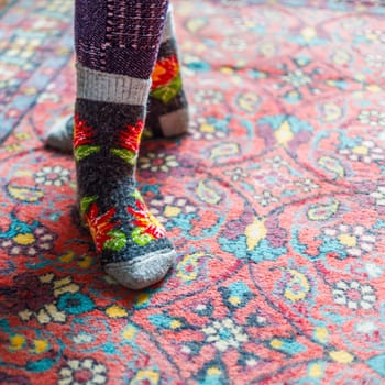 legs in warm woven wool socks standing on red ornate carpet floor at home backlit with a window light