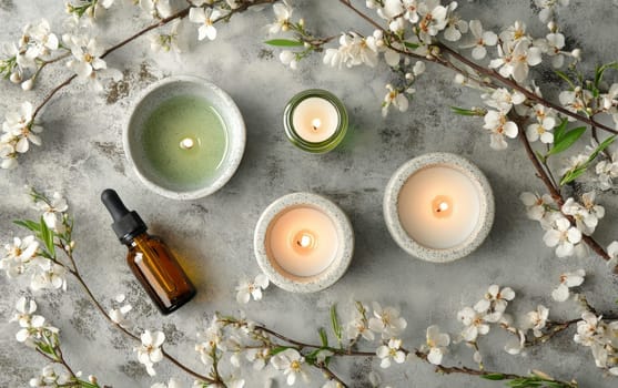 Tranquil spa composition with lit candles, essential oil bottle, and blooming cherry blossom branches on a textured gray background.