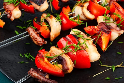 Grilled octopus skewers garnished with bell pepper. Seafood on a black plate.