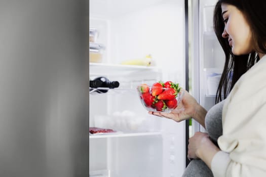 Thoughtful hungry young pregnant woman standing in front of open fridge, takes strawberry from refrigerator in middle of night at home in kitchen, closeup. Healthy eating, nutrition, pregnancy diet