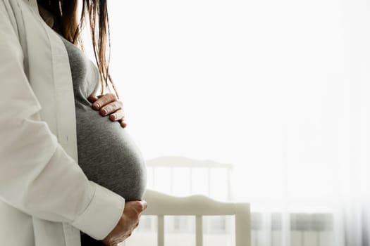 Mid-section portrait of pregnant woman or lady holding her baby bump or belly with hands. Standing near bed. Preparation for new family member. Expecting new life. Baby, new life concept. Last months