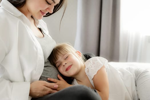 Mid-section portrait of happy, beautiful pregnant woman with cute little lovely daughter listening tummy, touching baby bump belly, girl hugging mother at home. Pregnancy, family, love concept