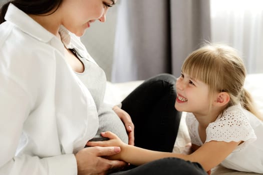 Midsection portrait of happy, beautiful, young careless pregnant woman, mother with cute little lovely daughter touching baby bump belly. Sitting together on bed at home, smiling each other. New life
