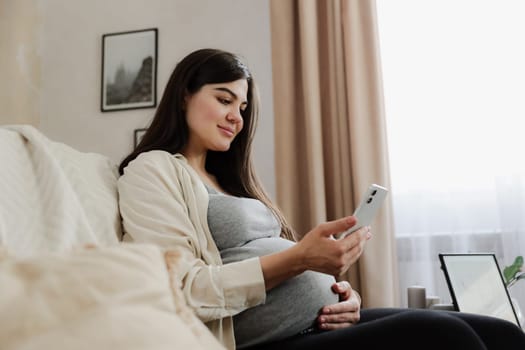 Beautiful smiling pregnant woman with baby belly use mobile cell phone, do online shopping, browse internet, watch podcast while sitting on sofa at home. Having doctor video call medical appointment