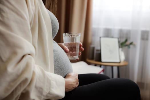 Close up of pregnant woman sitting on sofa at home, drinking, holding glass of water in hand. Pregnancy, prenatal health and wellness, healthy hydration. Medical, pharmacy and healthcare concept
