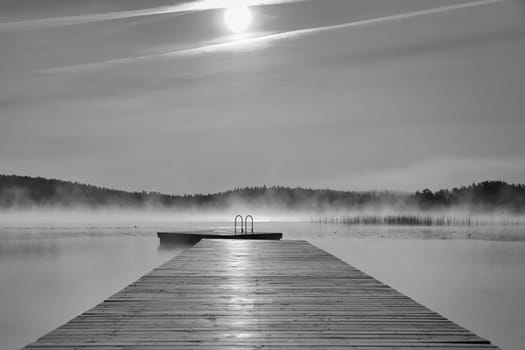 Wooden jetty in the morning at sunrise in the fog, on a Swedish lake in black and white. A floating island in the background. Scandinavian nature