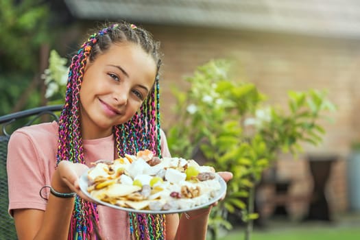 Portraiit  of cute young girl with colorful braids in her hair is holding a plate with cheese outside. Horizontally.