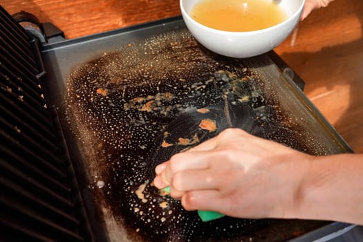 Cleaning electric grill dirty plate, detail on woman hand holding bowl with soup and sponge