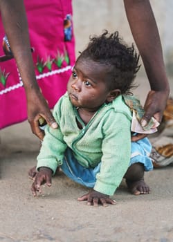 Ranohira, Madagascar - April 29, 2019: Unknown little Malagasy kid, barefoot, crawling on dusty footpath, mother helping her getting up. People in Madagascar are poor but cheerful