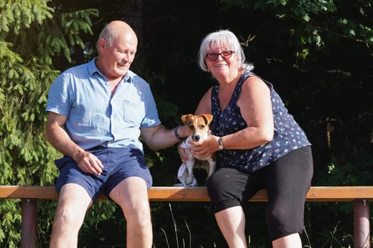 Senior couple sitting on wooden bench with small Jack Russell terrier dog, blurred trees in background