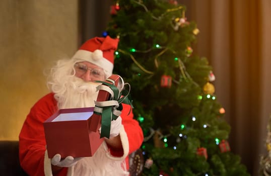 Santa Claus in red suit with hat holding an open red gift box with ribbon while sitting in front of decorated Christmas tree. Christmas celebration festive holiday concept.