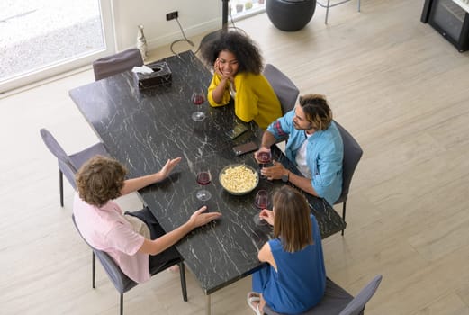 High angle view of multiethnic group of startup friends drinking red wine with popcorn and having conversation together at home.